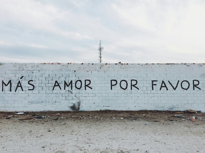 A painting on a white wall that reads MÁS AMOR POR FAVOR (more love please)